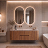 A modern floating bathroom vanity with a wood cabinet, integrated white countertop, and two oval backlit mirrors mounted on a textured beige wall. The scene includes brass faucets, decorative vases, and a partial white bathtub at right.