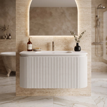 A modern floating bathroom vanity with a white ribbed cabinet and integrated sink, set against a warm beige stone wall with a lit arched mirror. A chrome faucet, soap dish, and small black vase with dried stems sit on the counter.